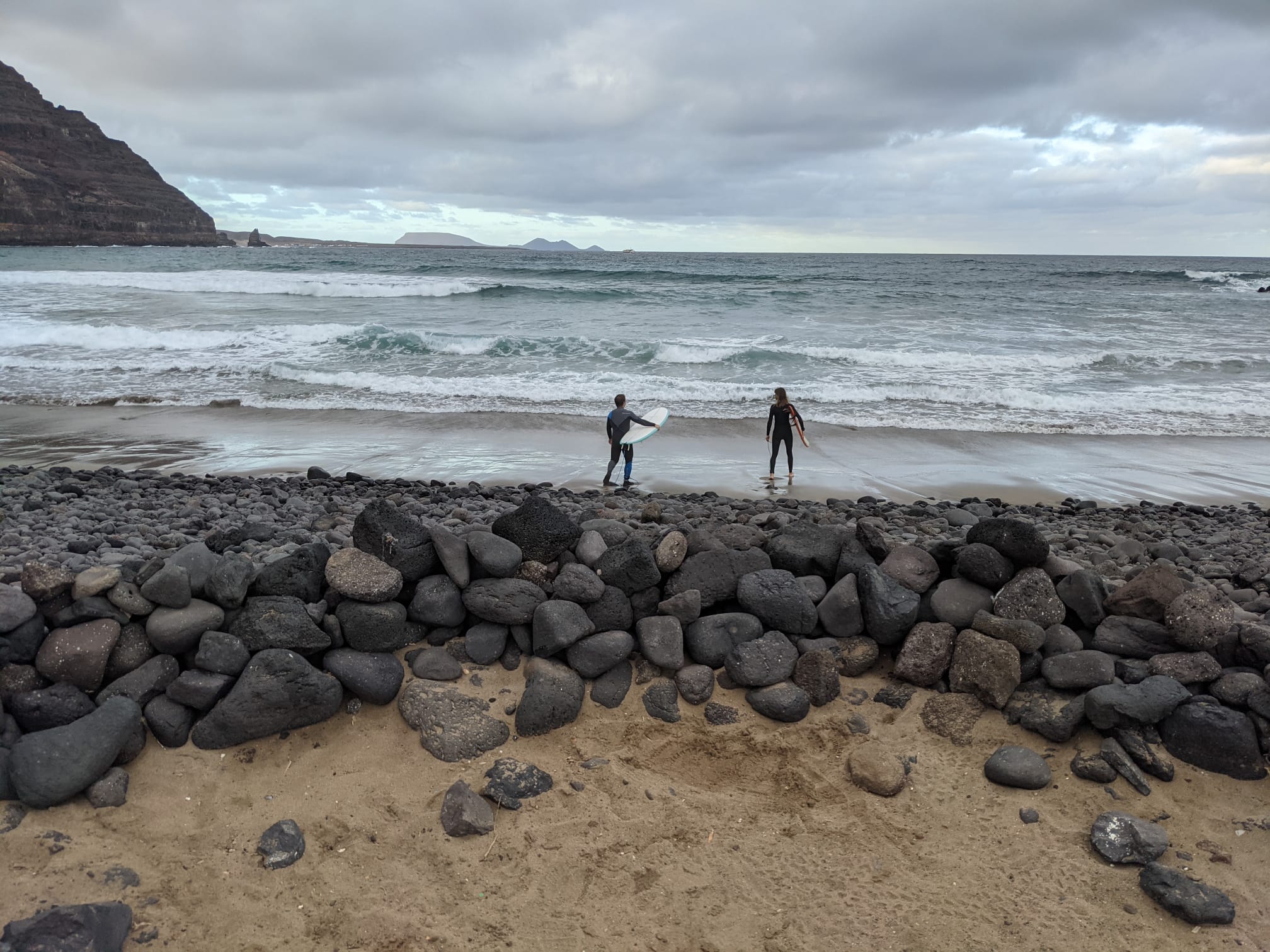 Two surfers in wetsuits, boards under their arms, standing at the water's edge on a sand and lava-rock beach, looking out at the waves